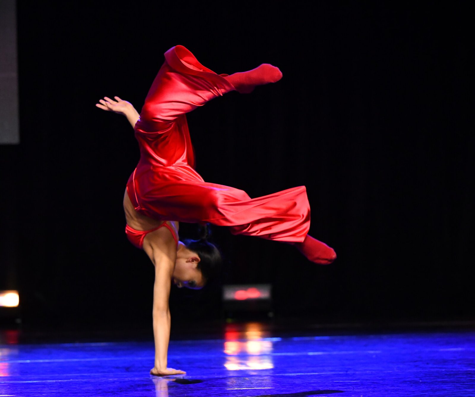 Glee Dango performing a one-handed handstand in a red costume on stage